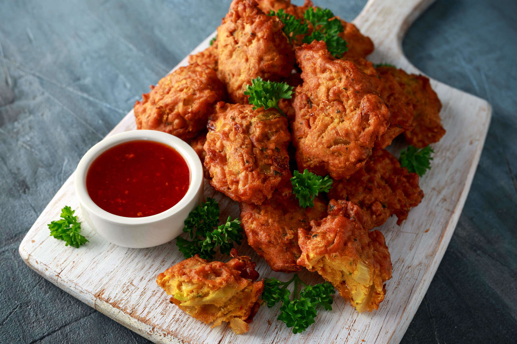 a stack of vegetable pakoras on a wooden board with chilli dipping sauce garnished with green herbs