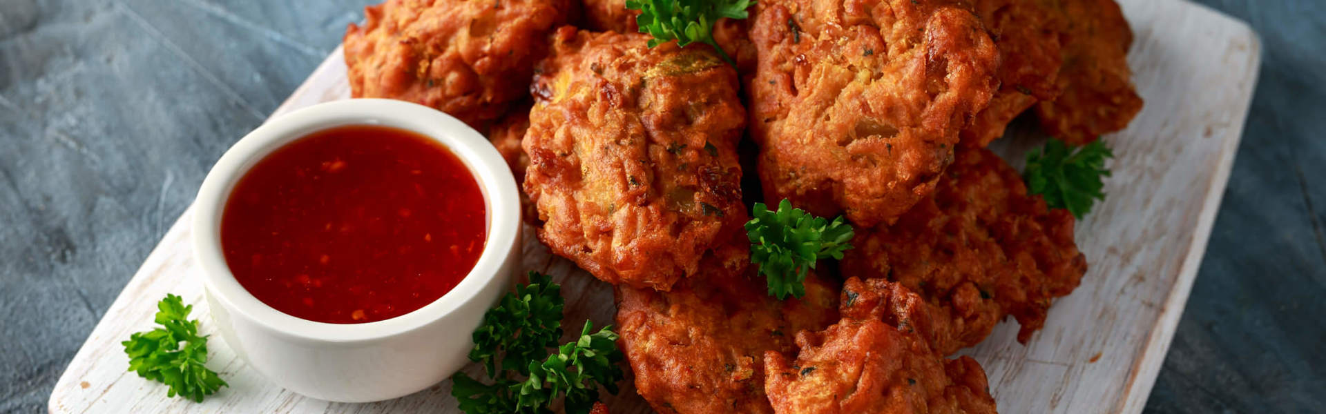a stack of vegetable pakoras on a wooden board with chilli dipping sauce garnished with green herbs