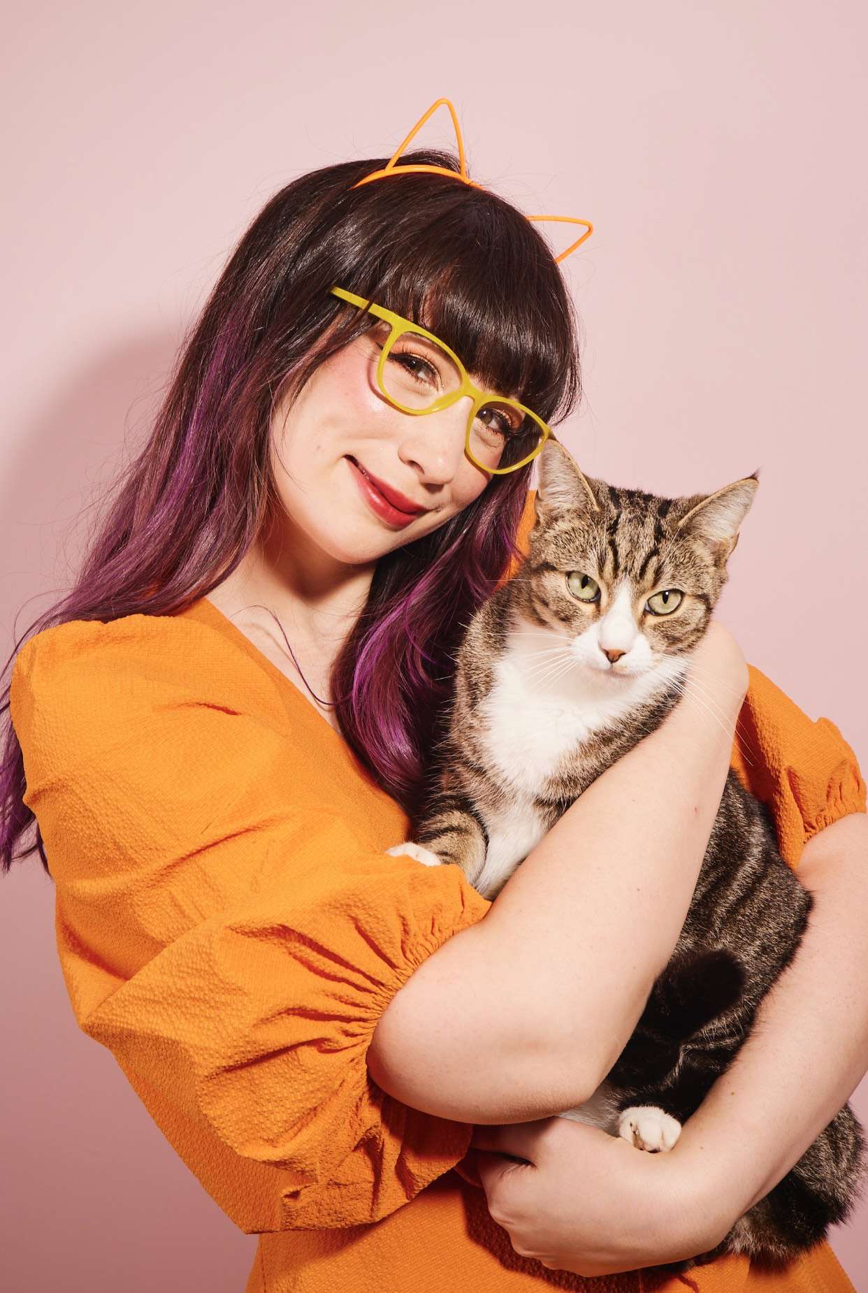 Main image: a photo of Kim Joy Author Of Ordinary Joy, cuddling her cat, wearing an orange top in front of a pink background.