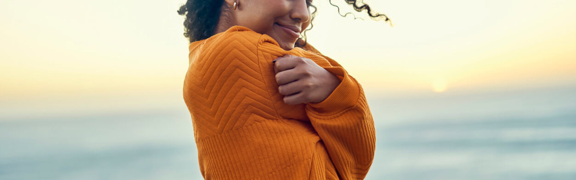 a woman wearing an orange jumper giving herself a hug with the beach horizon in the background