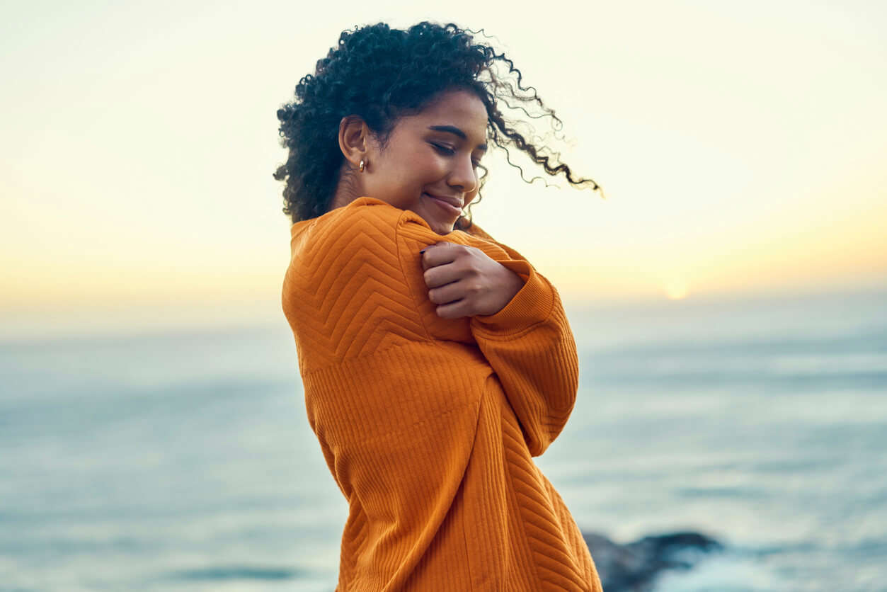 a woman wearing an orange jumper giving herself a hug with the beach horizon in the background