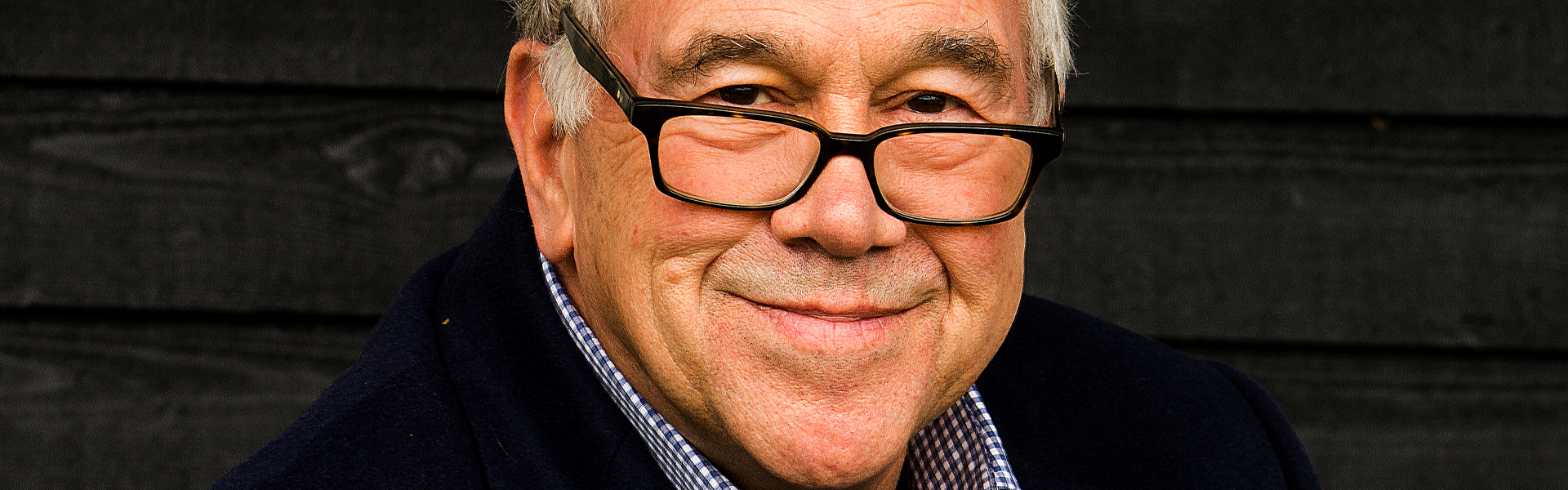 Main Image: a photo of cosy crime author Simon Brett, wearing a navy suit jacket, smiling at the camera.