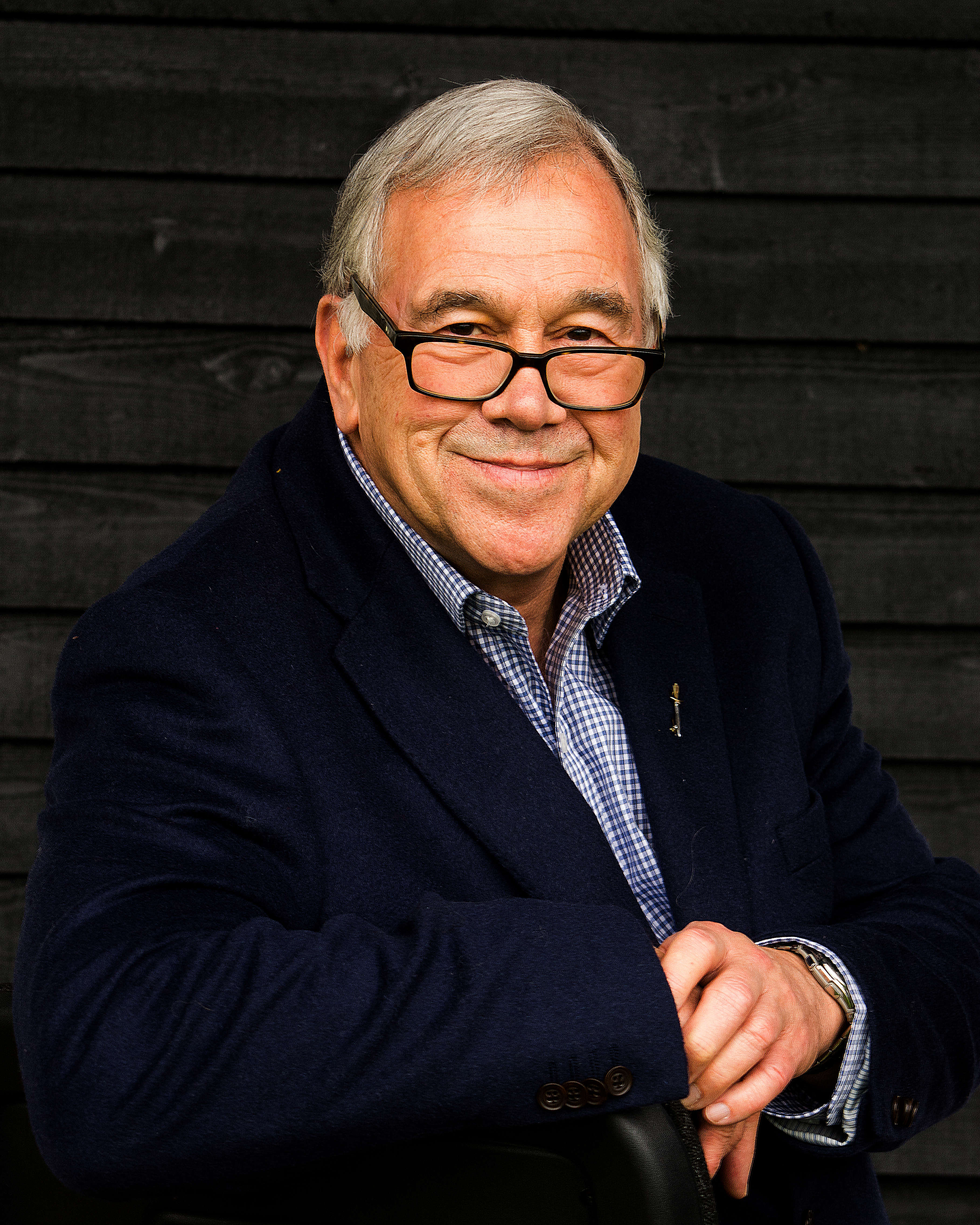 Main Image: a photo of cosy crime author Simon Brett, wearing a navy suit jacket, smiling at the camera.