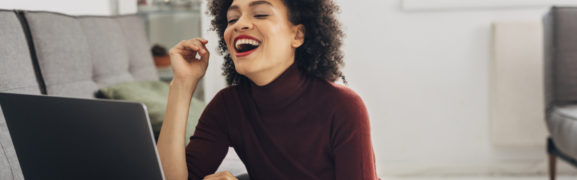 A woman in a red high neck jumper sat on the living room floor laughing whilst looking at her laptop