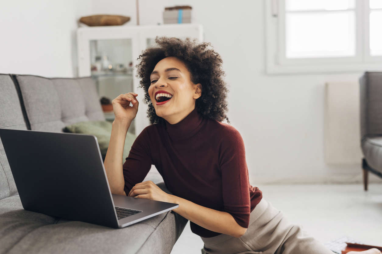 A woman in a red high neck jumper sat on the living room floor laughing whilst looking at her laptop