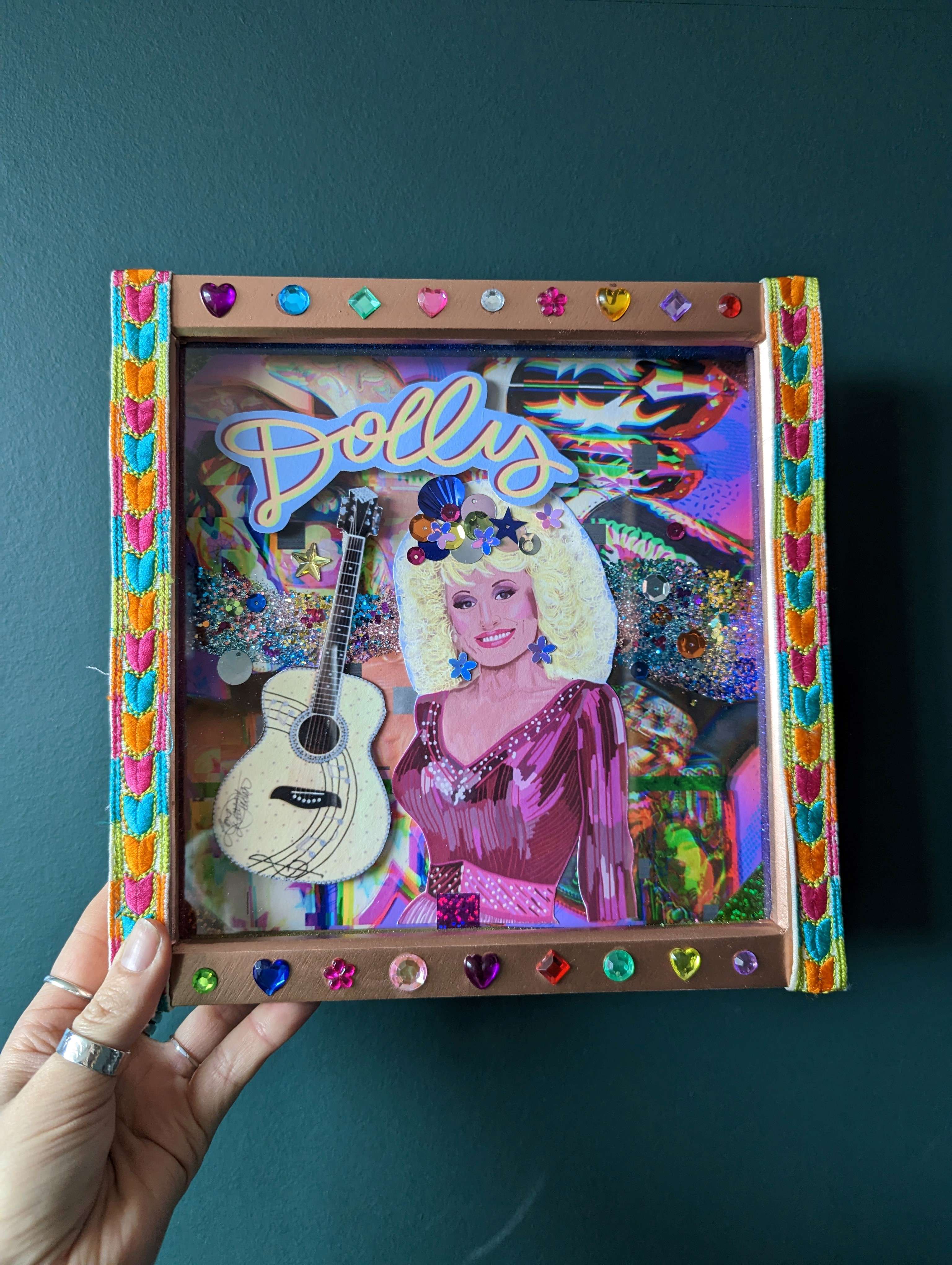 Multi-coloured shadow box shrine made with a variety of materials displaying a paper image of the singer Dolly Parton with the word "Dolly" displayed to the left of her