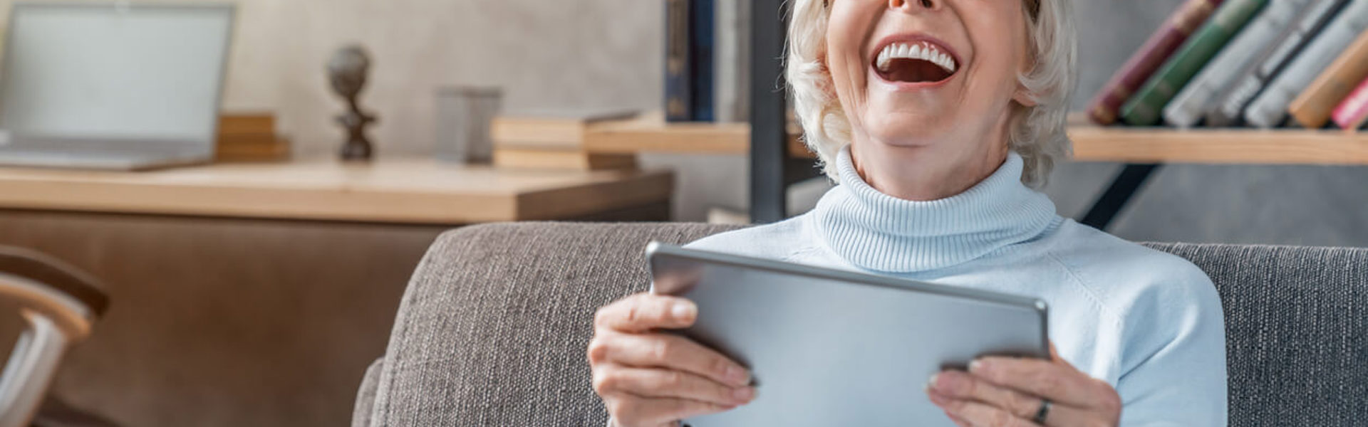 An older woman wearing glasses sat on a sofa holding a tablet and laughing