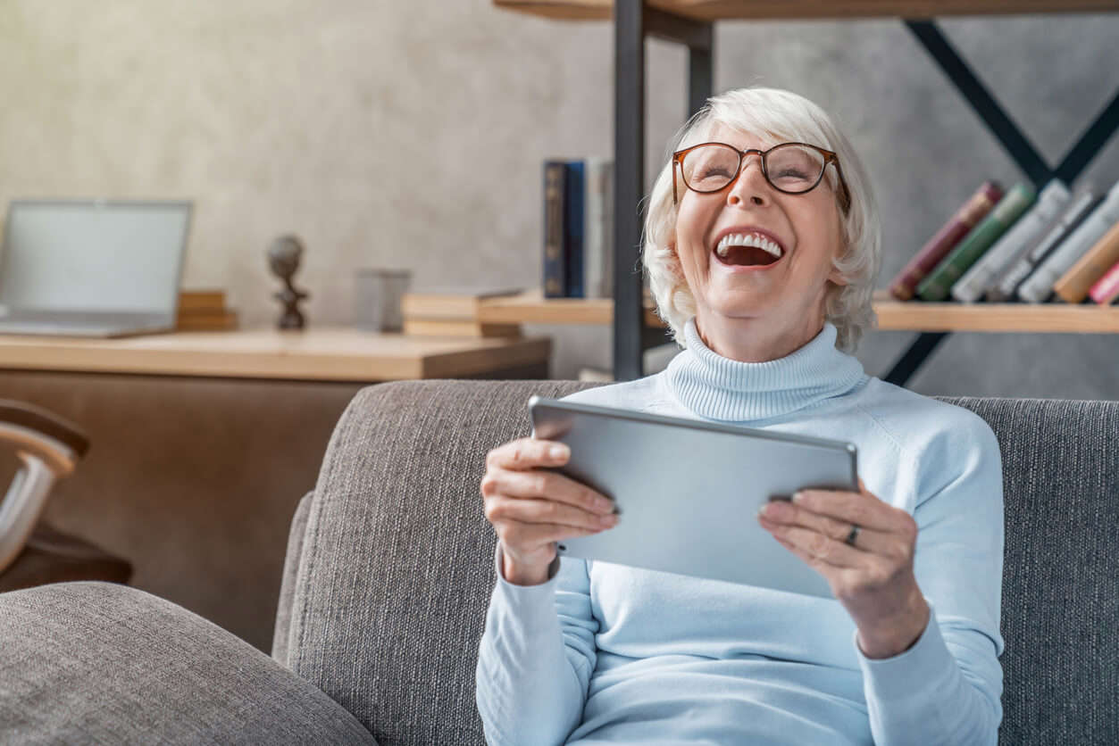 An older woman wearing glasses sat on a sofa holding a tablet and laughing
