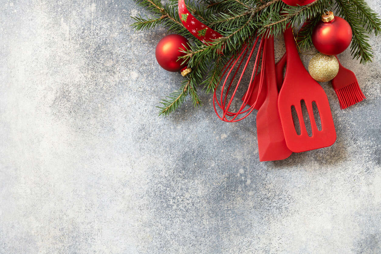 Red Christmas themed cooking utensils in a wreath with red and gold baubles on a grey background