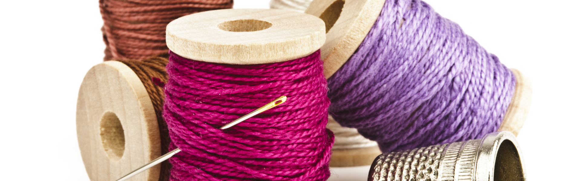 Main Image: An assortment of pink, purple and orange thread spools with a sewing needle and thimble.