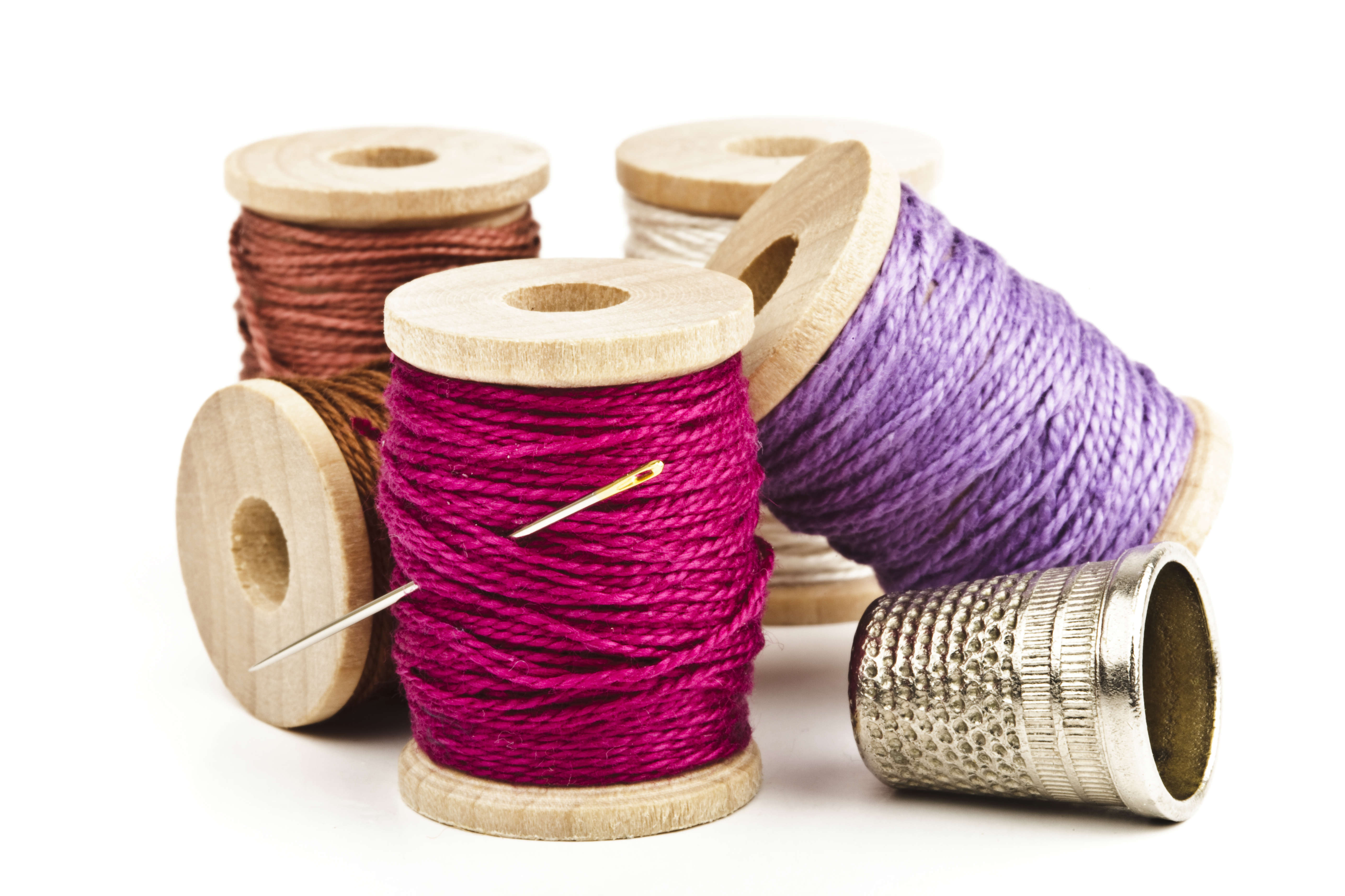 Main Image: An assortment of pink, purple and orange thread spools with a sewing needle and thimble.