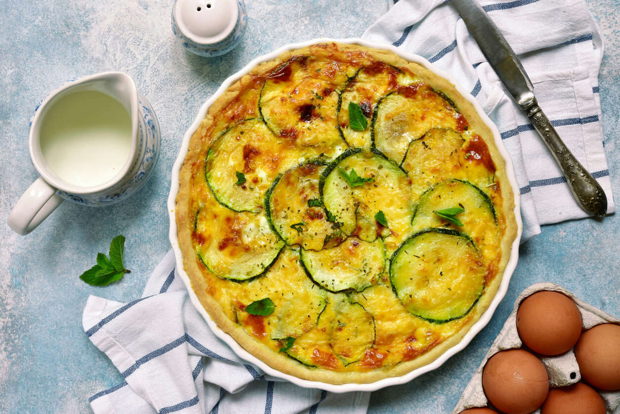 Mediterranean vegetables and hash brown quiche in an oven dish, on a towel with ingredients around it.