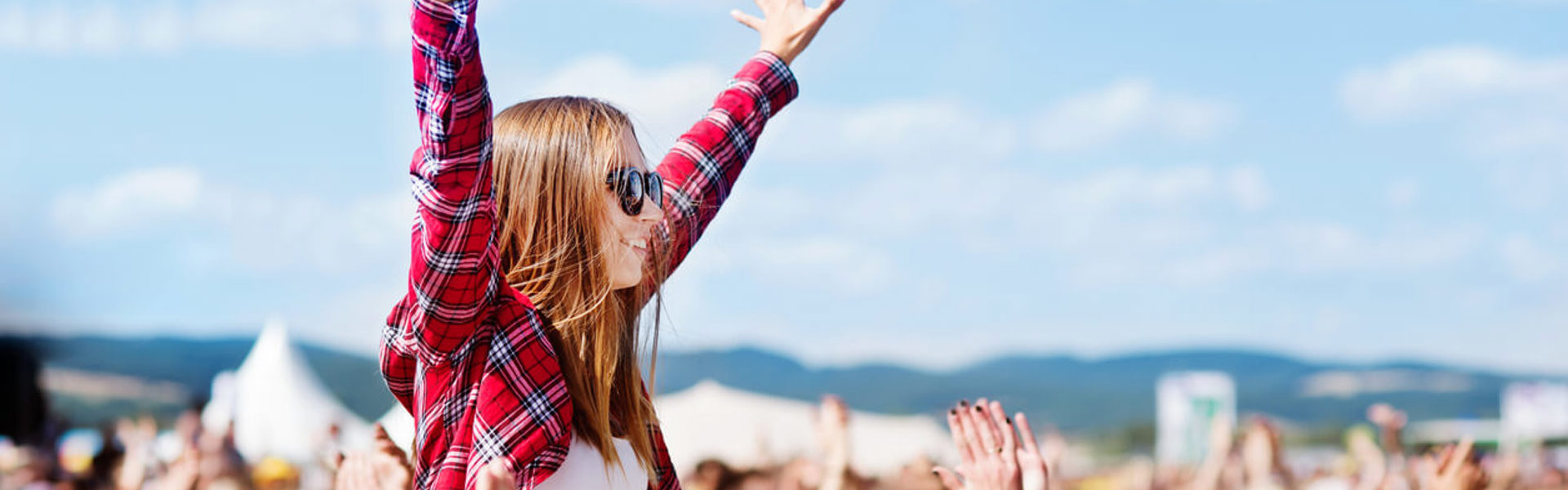 a woman wearing a red check shirt and sunglasses sitting on a friends shoulders at a festival with her hands in the air watching a concert