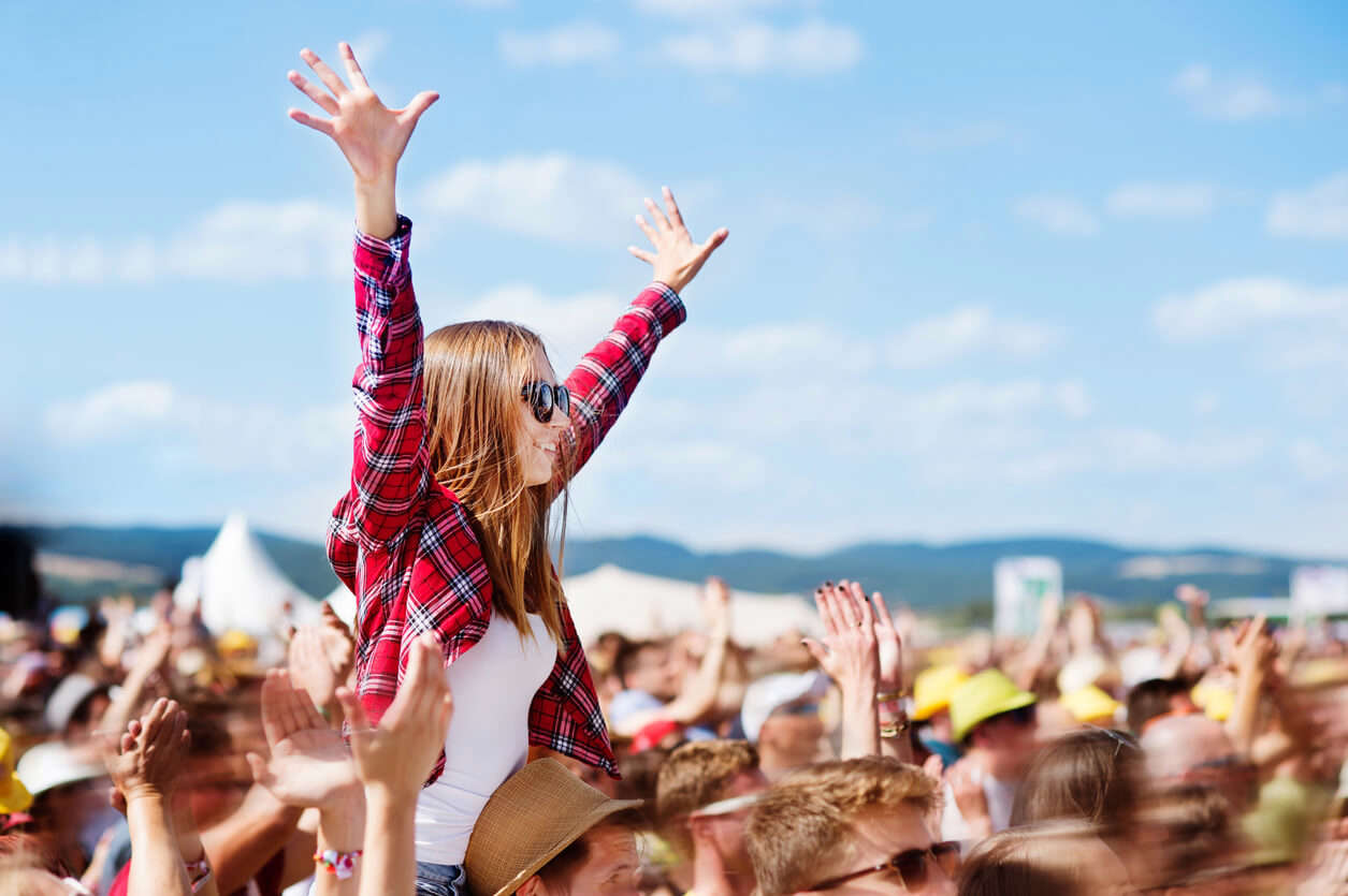 a woman wearing a red check shirt and sunglasses sitting on a friends shoulders at a festival with her hands in the air watching a concert