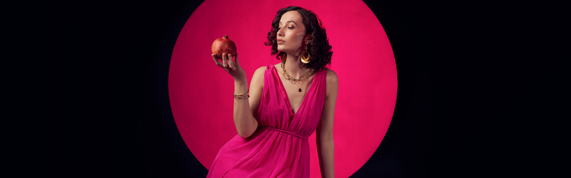 Main image: best selling YA Fantasy author Bea Fitzgerald in a beautiful bright pink floor length dress, holding a pomegranate looking at it. With a black background and pink circle behind her. 
