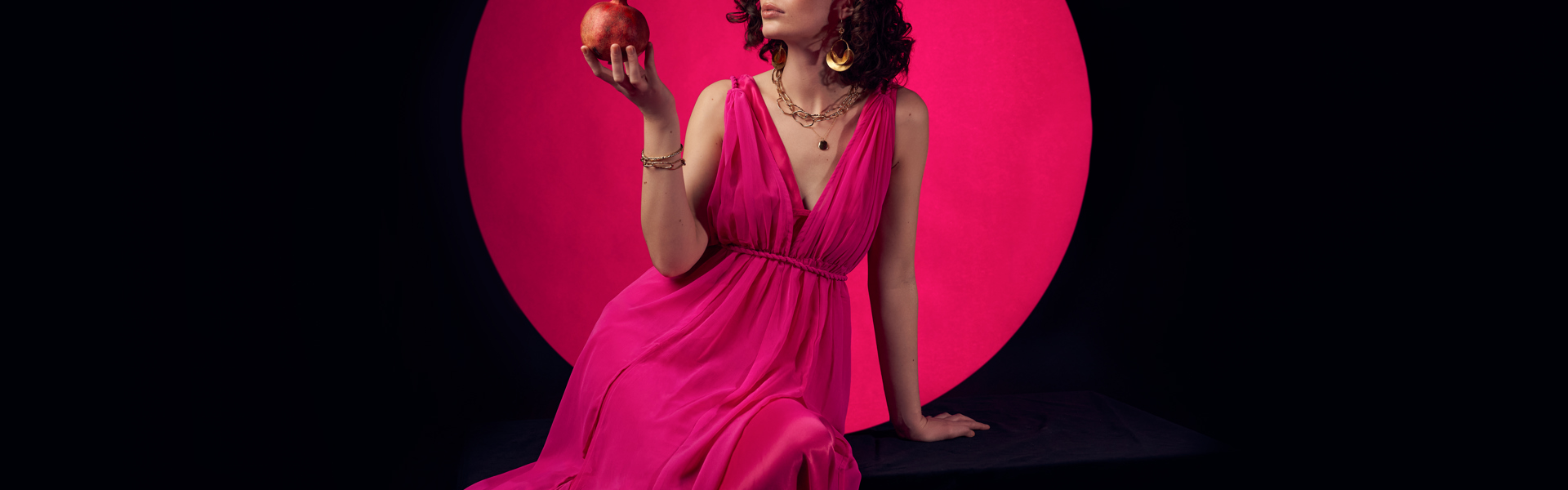 Main image: best selling YA Fantasy author Bea Fitzgerald in a beautiful bright pink floor length dress, holding a pomegranate looking at it. With a black background and pink circle behind her. 