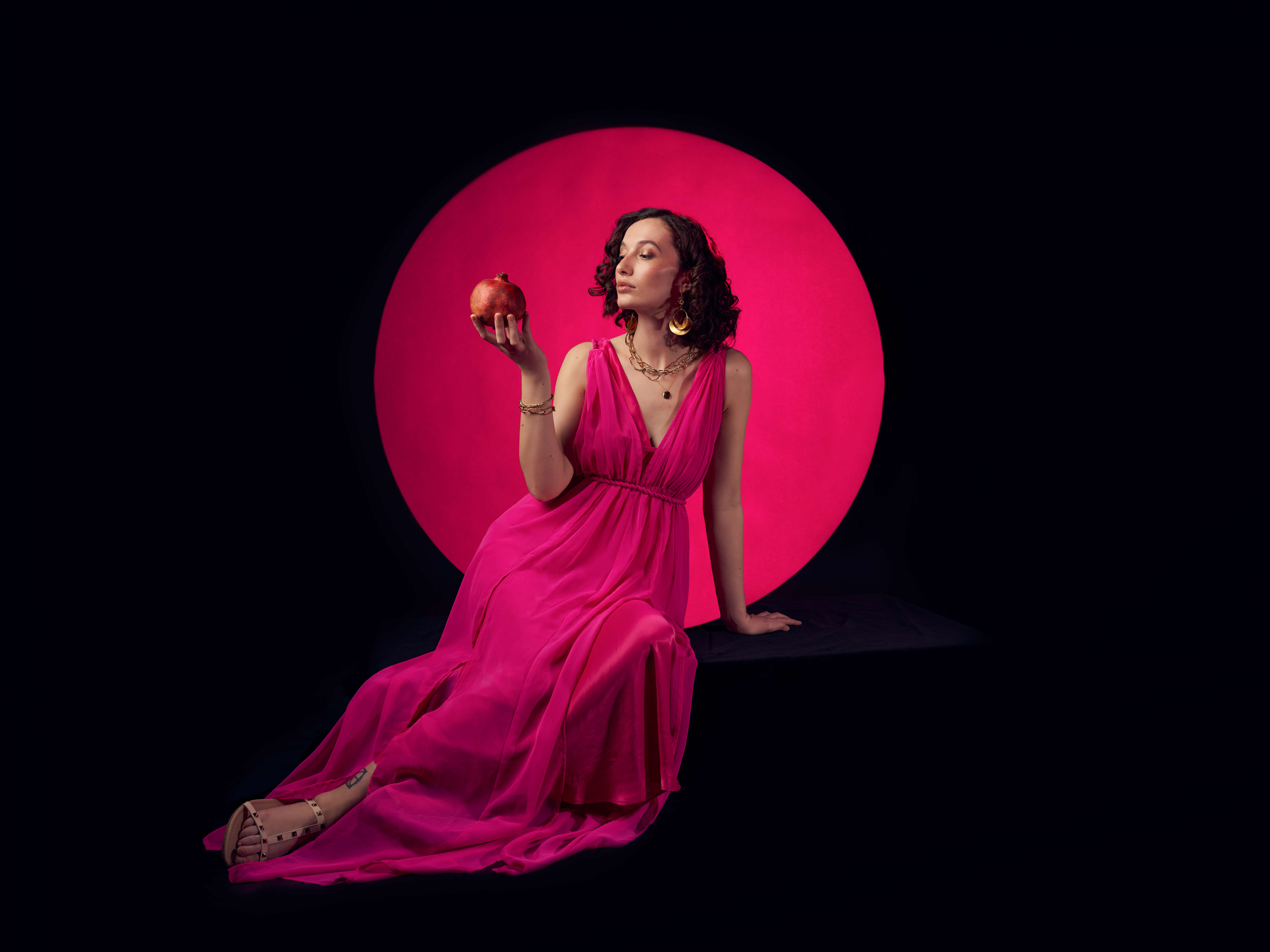 Main image: best selling YA Fantasy author Bea Fitzgerald in a beautiful bright pink floor length dress, holding a pomegranate looking at it. With a black background and pink circle behind her. 