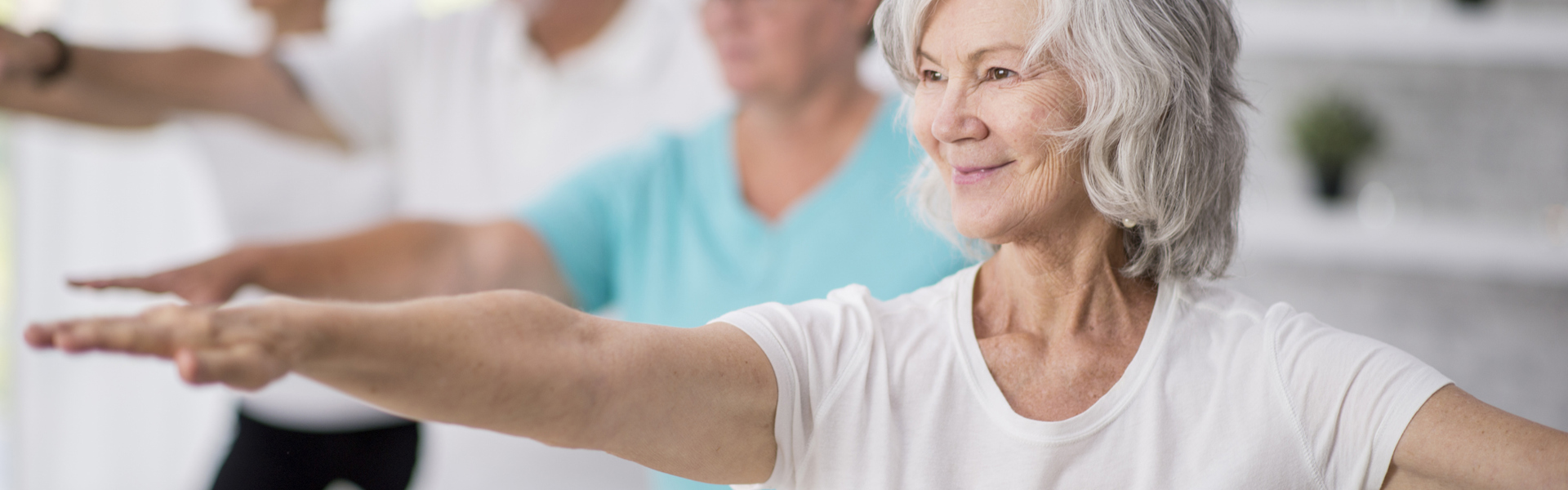Older adults practising Balance exercises for falls prevention, standing with their arms out in a yoga pose. The woman in focus at the front is smiling, with short grey hair and a white t-shirt.