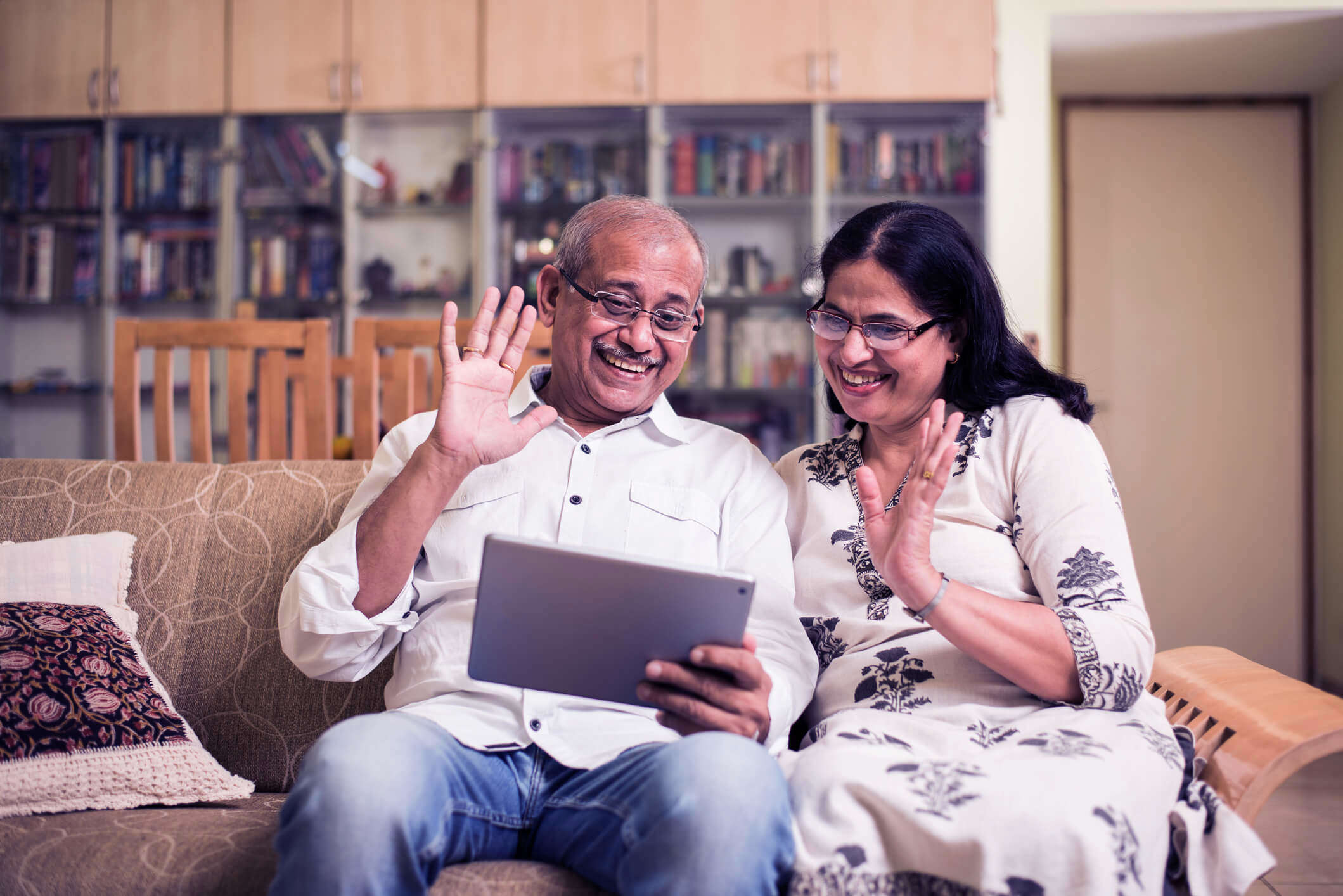 an older couple wearing glasses sat on a sofa and waving at an ipad