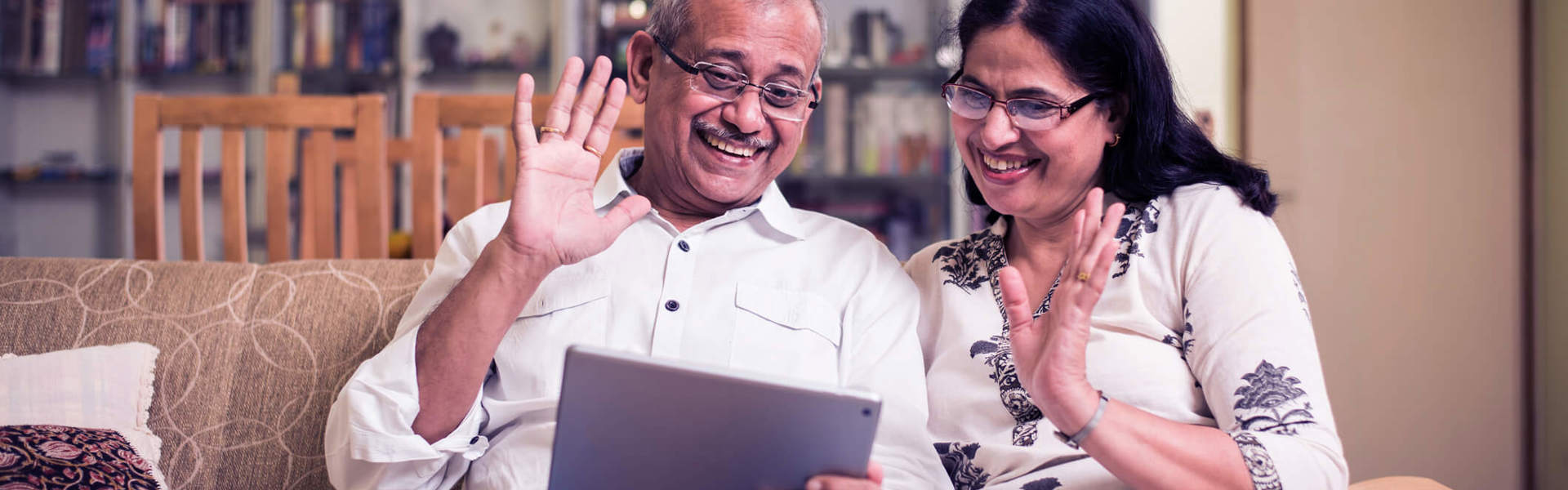 an older couple wearing glasses sat on a sofa and waving at an ipad