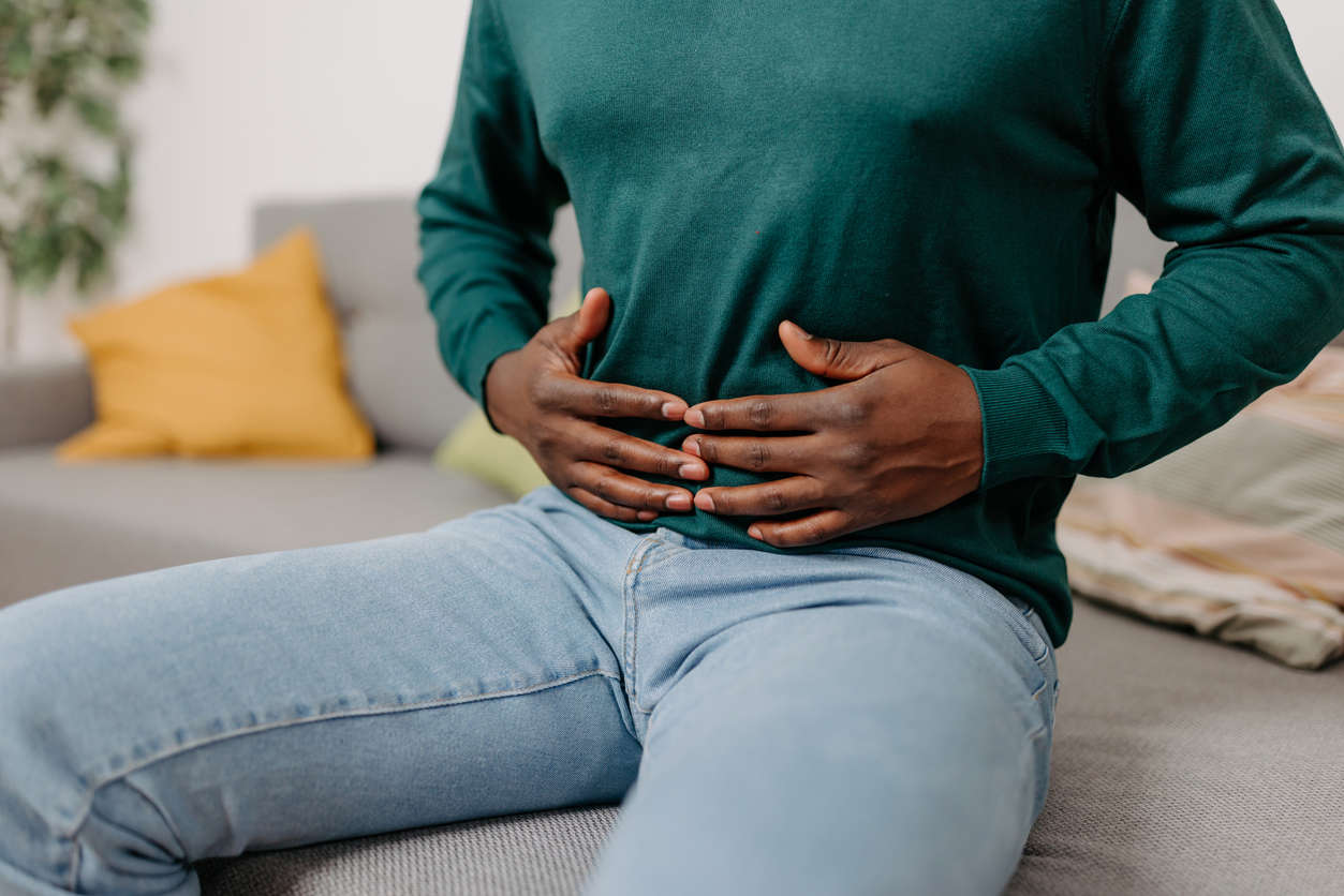 a person in a dark green top and blue jeans holding their stomach whilst sat on a sofa