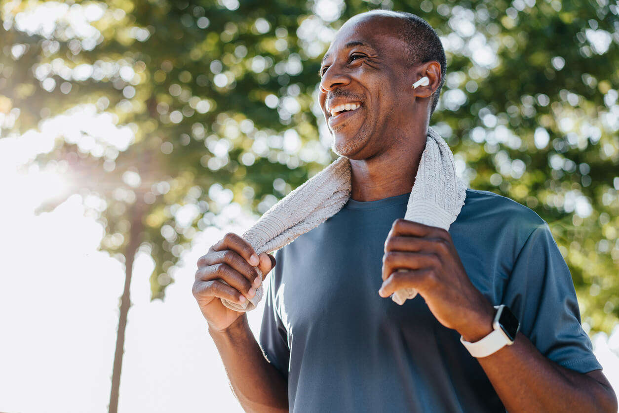 A middle aged man wearing wireless headphones exercising outdoors and smiling with a work out towel 