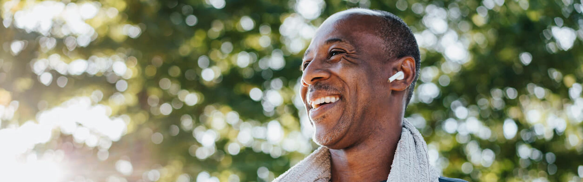 A middle aged man wearing wireless headphones exercising outdoors and smiling with a work out towel