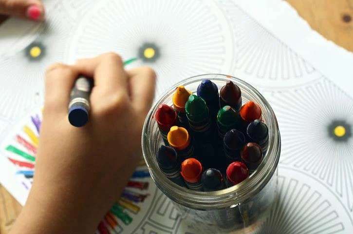 a hand holding a crayon and drawing on paper with a pot of multicoloured crayons next to it