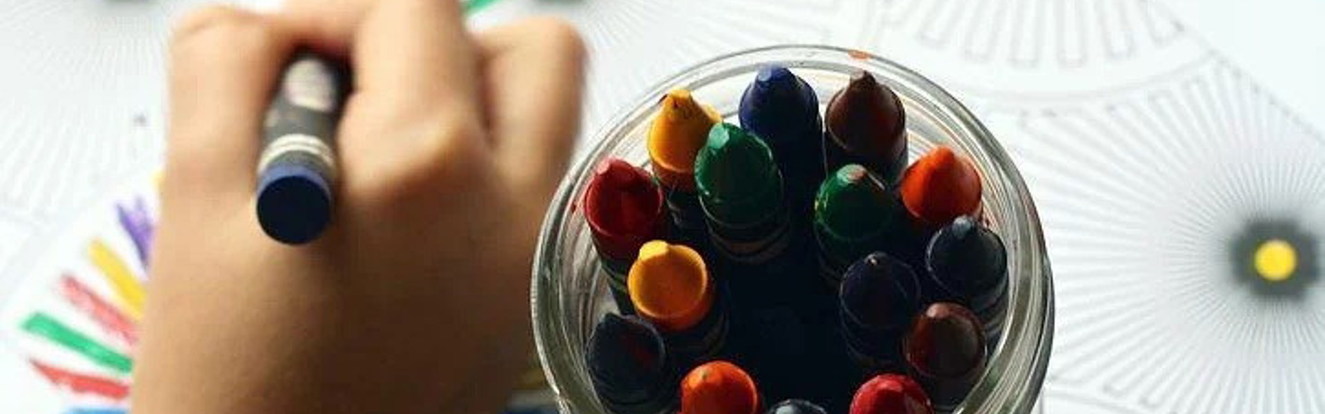 a hand holding a crayon and drawing on paper with a pot of multicoloured crayons next to it