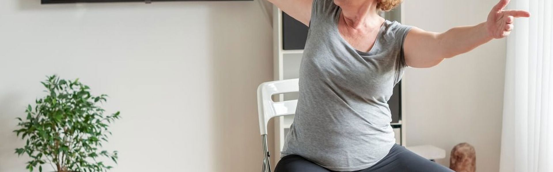 Main Image: Woman sitting in her living room doing chair yoga stretches
