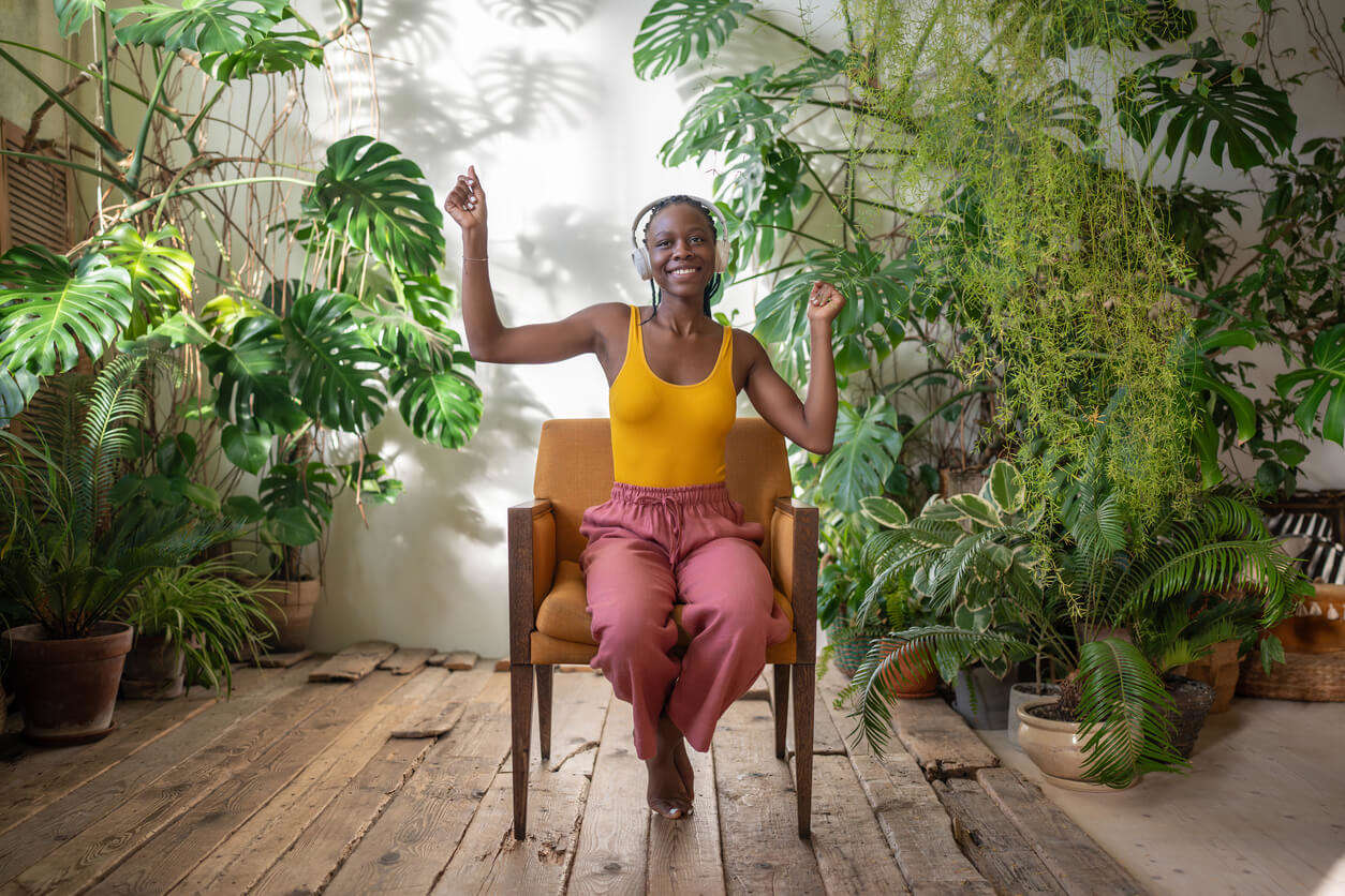 A woman wearing a yellow t-shirt and pink trousers sat on a wooden chair dancing surrounded by plants