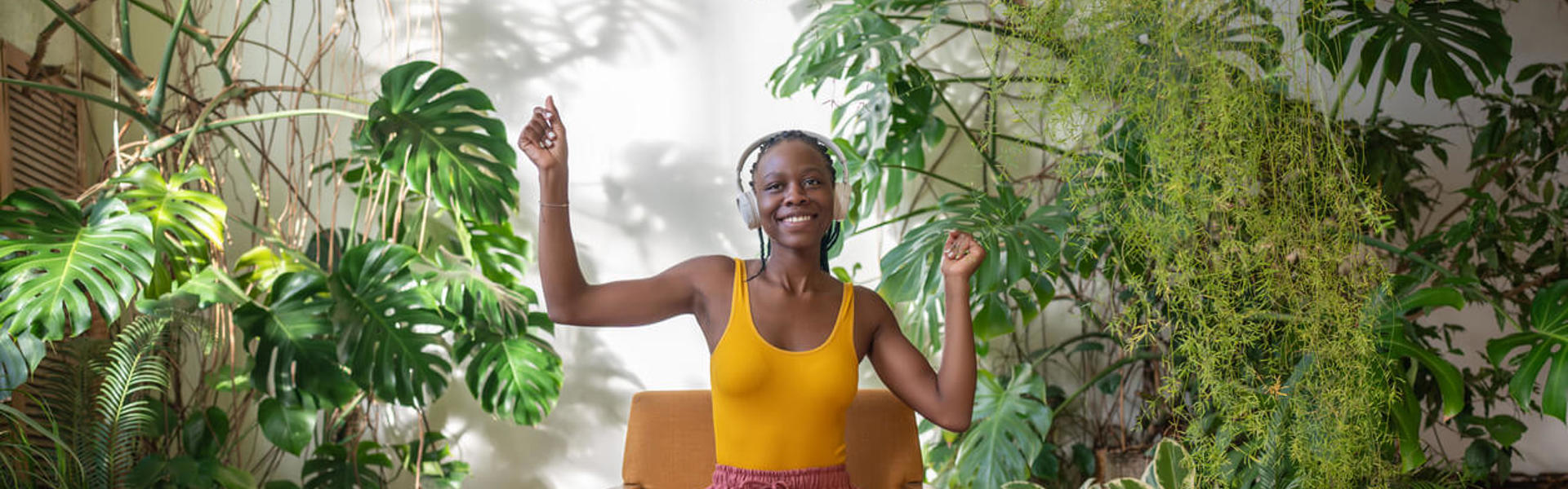 A woman wearing a yellow t-shirt and pink trousers sat on a wooden chair dancing surrounded by plants