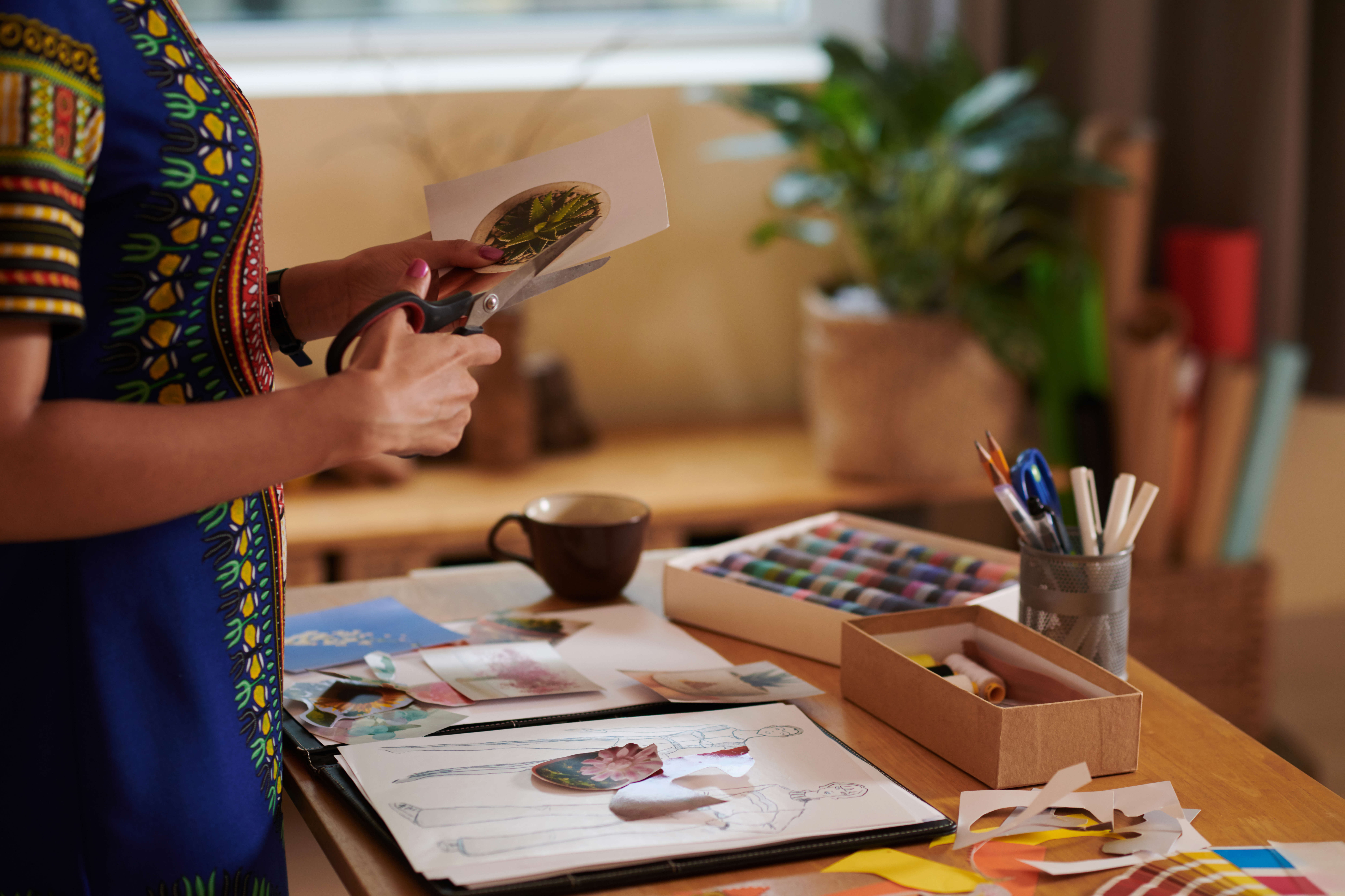 A person making a collage art picture using scissors and scraps of paper and magazines on a wooden table surrounded by art materials