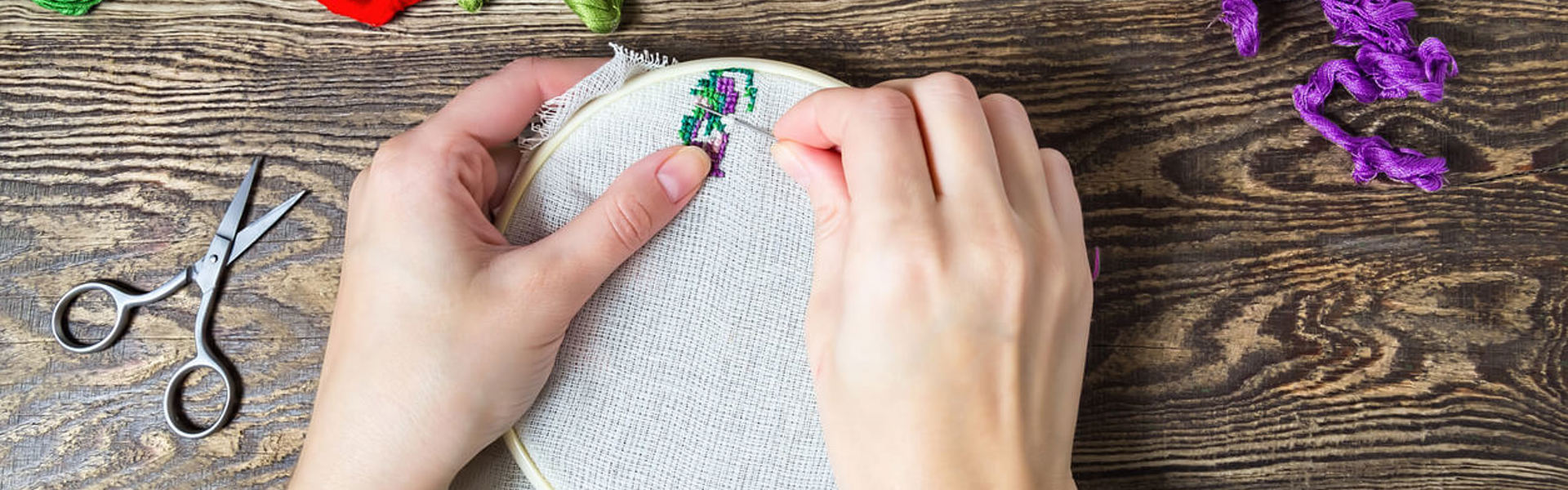 a close up of a pair of hands using an embroidery hoop to embroider purply and green flowers with gold green red and purple thread next to it