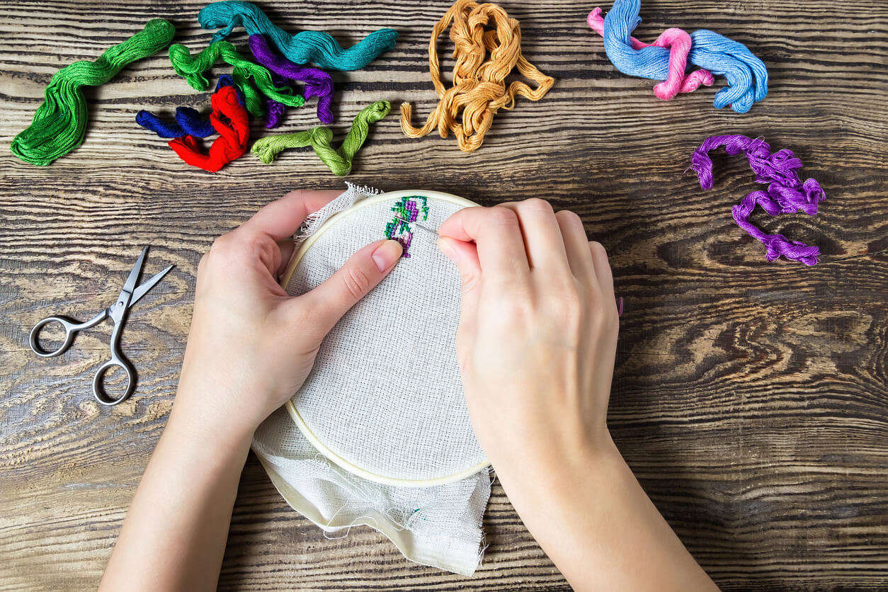 a close up of a pair of hands using an embroidery hoop to embroider purply and green flowers with gold green red and purple thread next to it