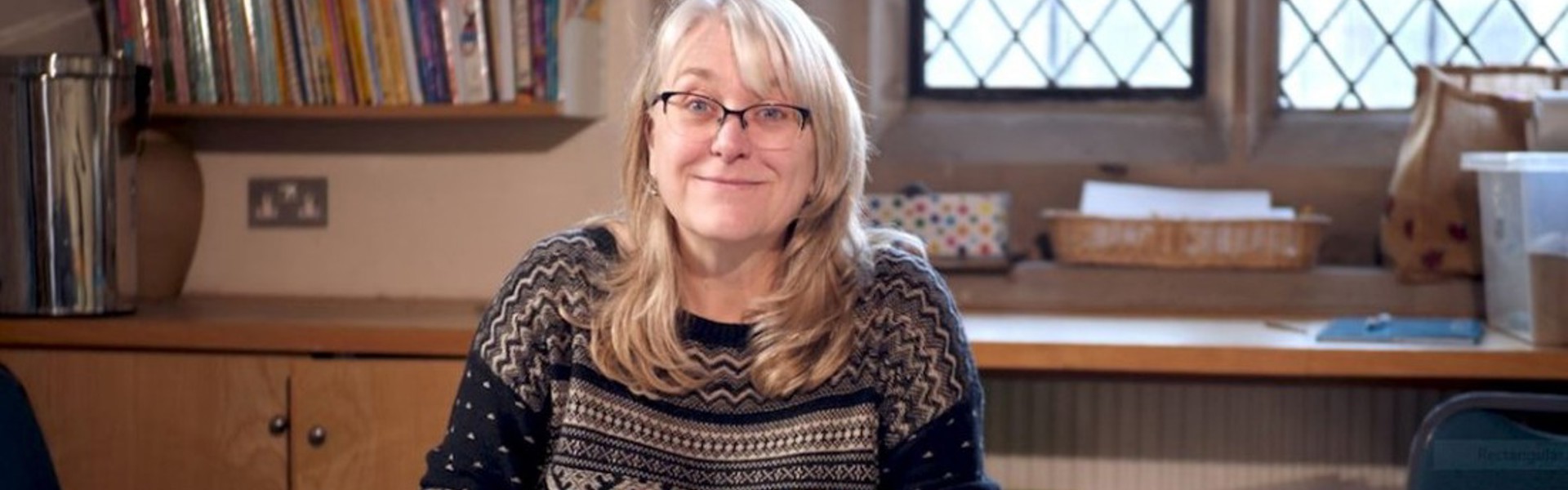 Main image: Alison Mott sat writing at her desk, smiling wearing a navy jumper.
