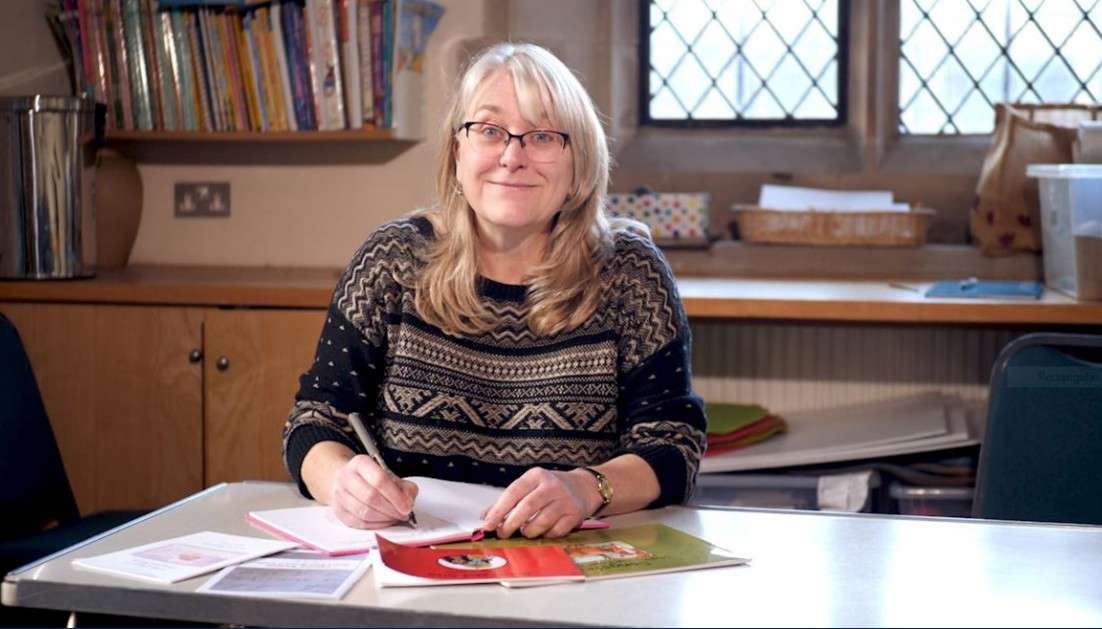 Main image: Alison Mott sat writing at her desk, smiling wearing a navy jumper.