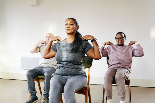 Main image: A group of three people taking part in a Gentle Chair Yoga class, sat in chairs with feet flat on the floor and hands touching their shoulders, smiling.