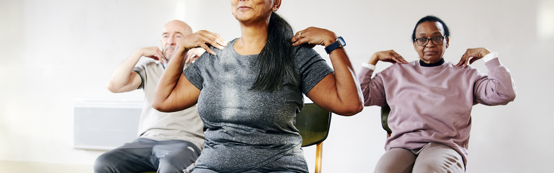 Main image: A group of three people taking part in a Gentle Chair Yoga class, sat in chairs with feet flat on the floor and hands touching their shoulders, smiling.