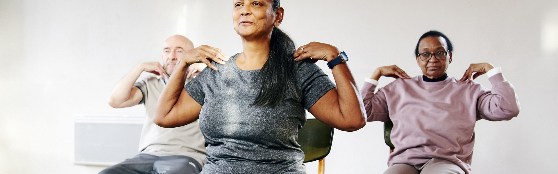 Main image: A group of three people taking part in a Gentle Chair Yoga class, sat in chairs with feet flat on the floor and hands touching their shoulders, smiling.