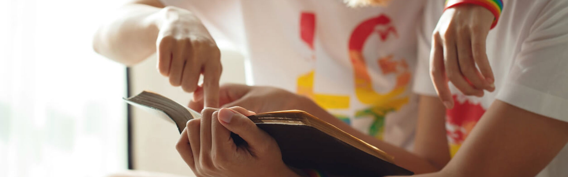 Young women LGBT sitting on sofa reading book together, both wearing rainbow wristbands.