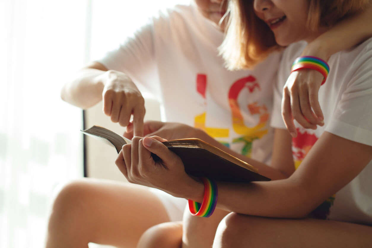 Young women LGBT sitting on sofa reading book together, both wearing rainbow wristbands.