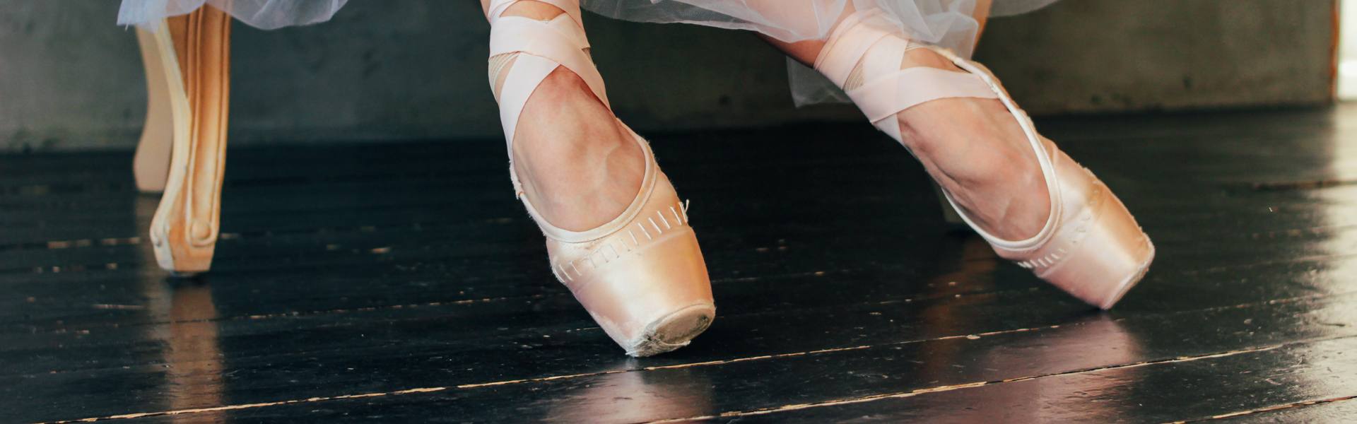 Main image: a close up of a ballet dancer sat in a chair, in their pale peach ballet shoes, wearing a long white tutu.