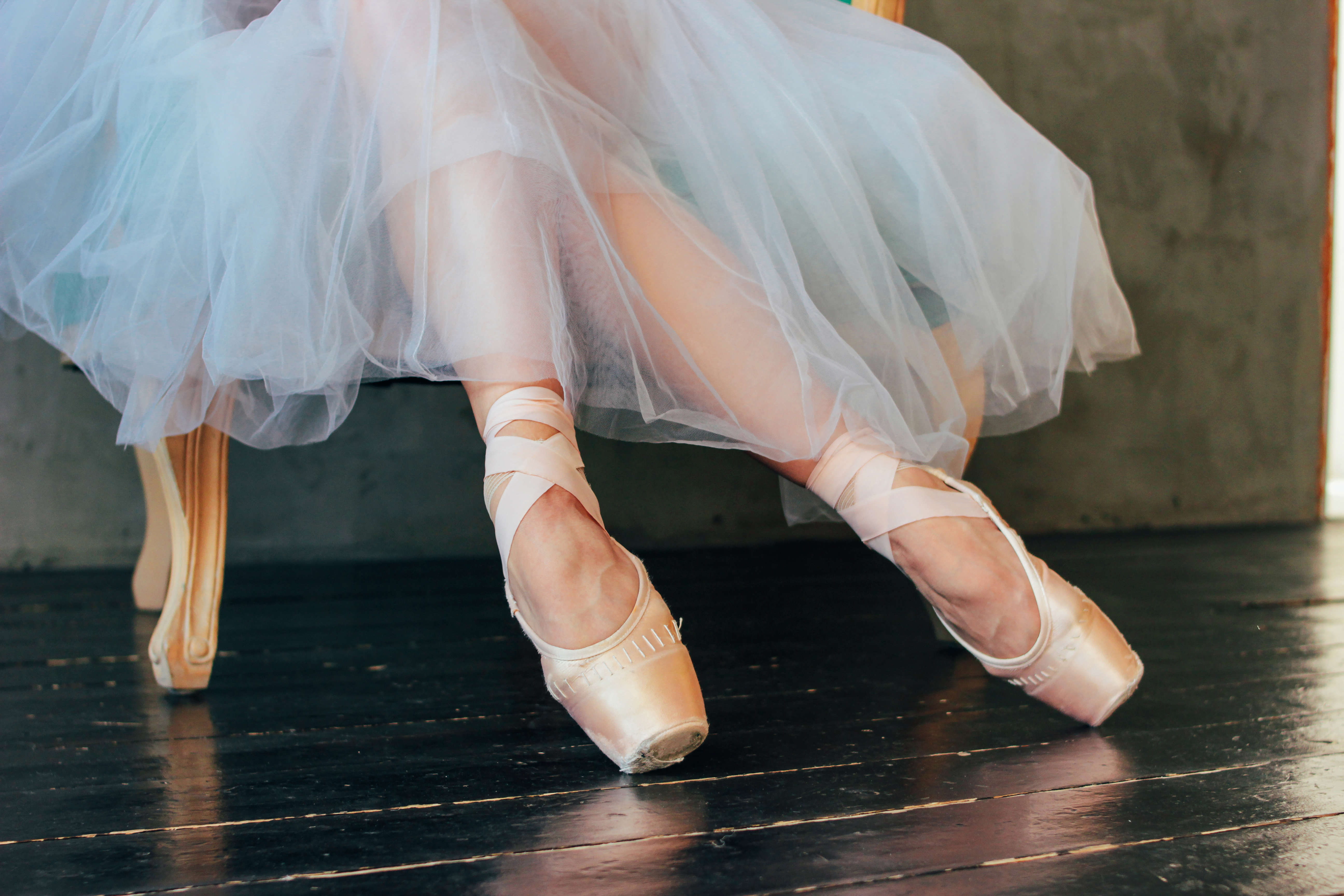 Main image: a close up of a ballet dancer sat in a chair, in their pale peach ballet shoes, wearing a long white tutu.