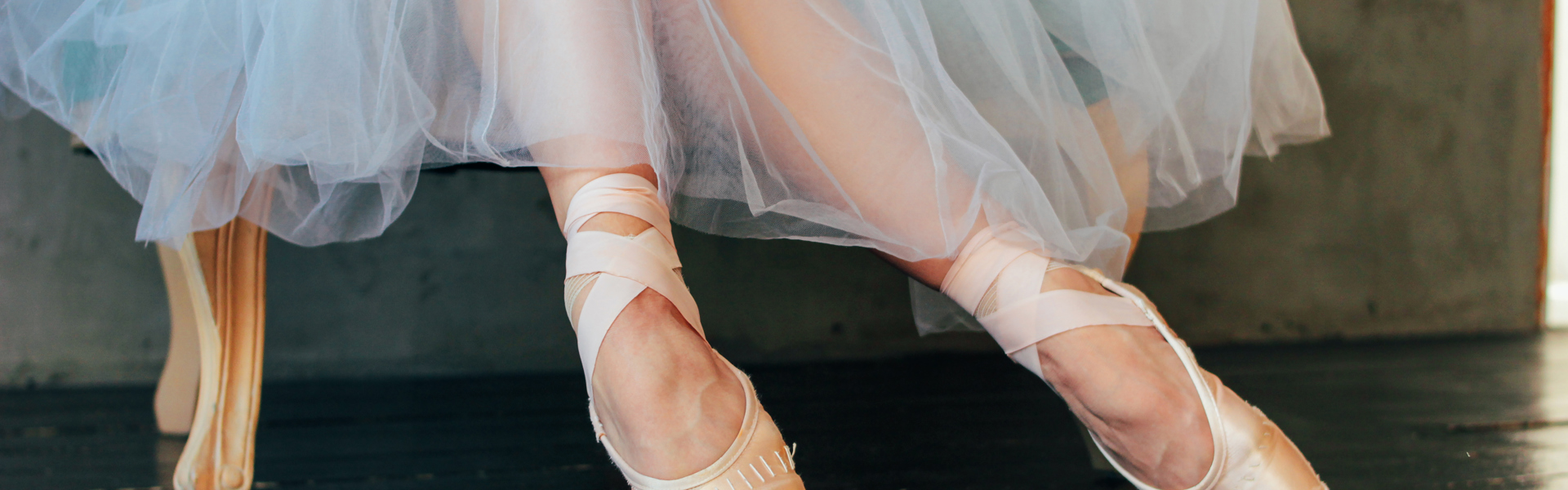 Main image: a close up of a ballet dancer sat in a chair, in their pale peach ballet shoes, wearing a long white tutu.