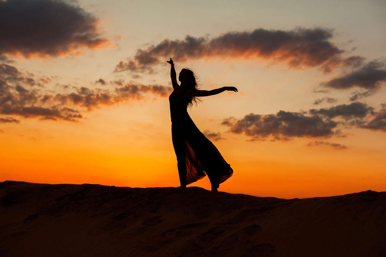 a silhouette of a woman with long hair dancing on a orange horizon in a skirt