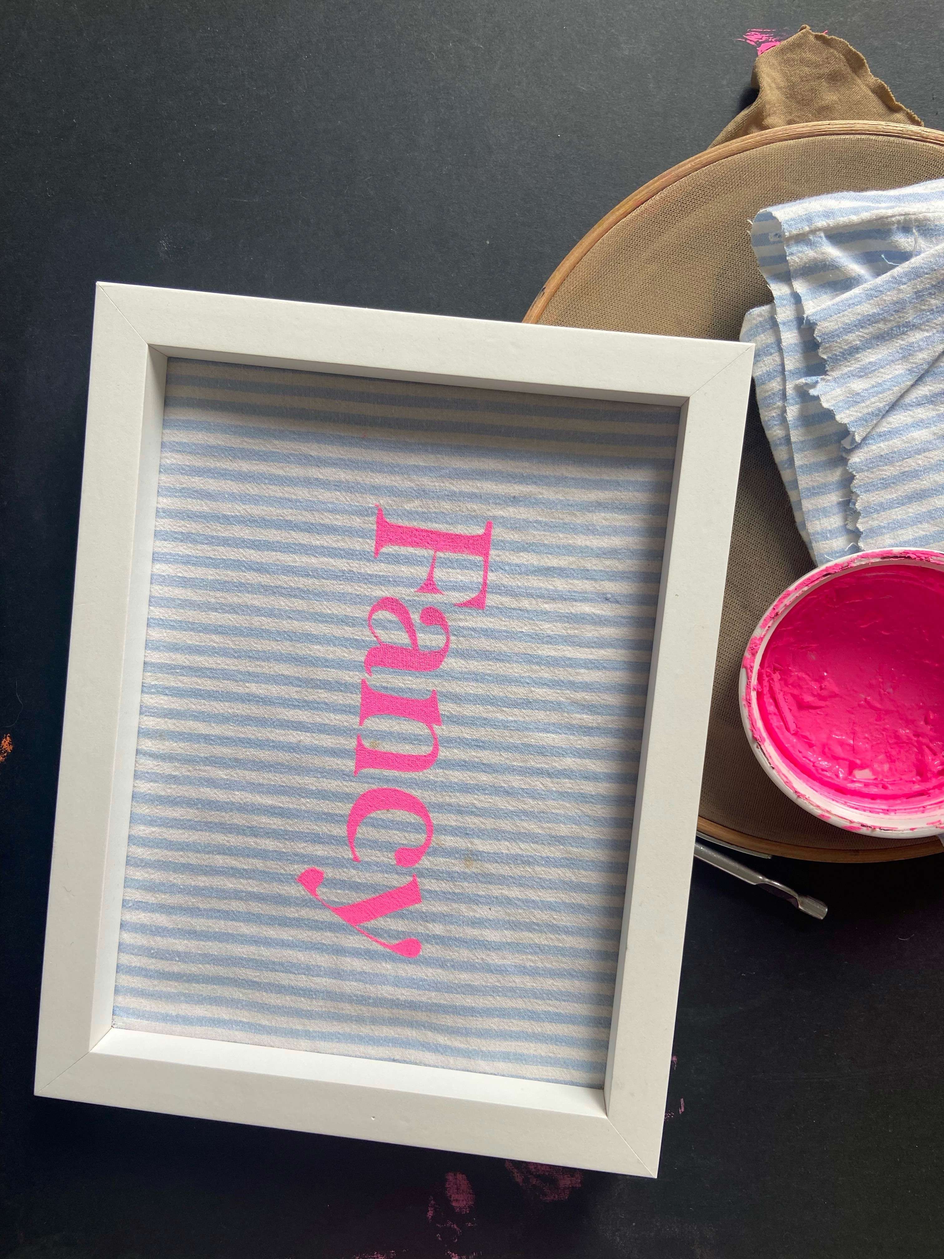 a striped screen print work with neon pink text printed onto it and framed in a white frame 