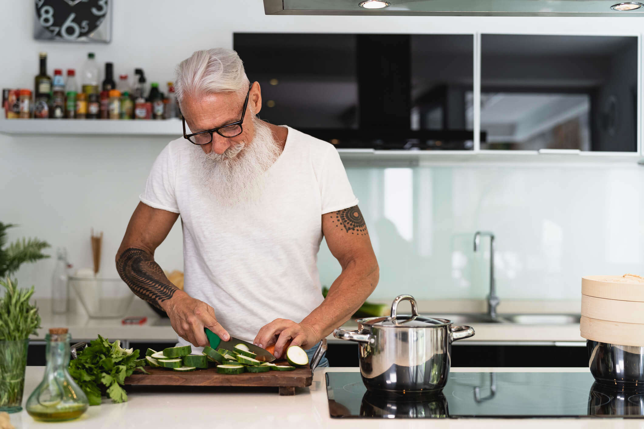 a man with a beard and glasses in a white t-shirt slicing courgette in a kitchen