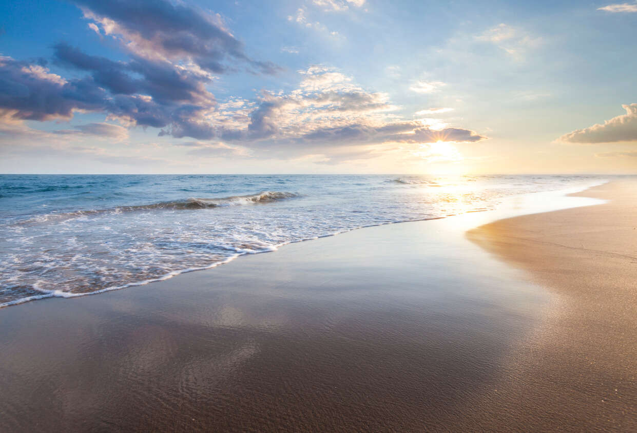 a landscape picture of waves on a sandy beach with a sunset in the background