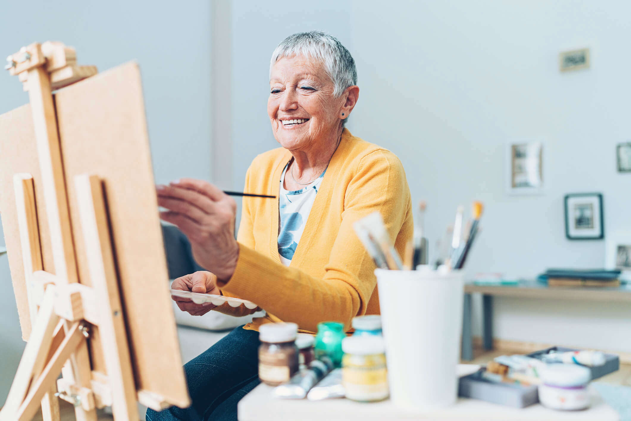 An older woman wearing a yellow cardigan sat at a table painting using a table top easel